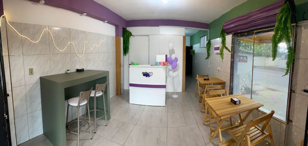 Visão interna do balcão de atendimento com as máquinas de cartão e mesas na área externa do Ateliê do Açaí.
